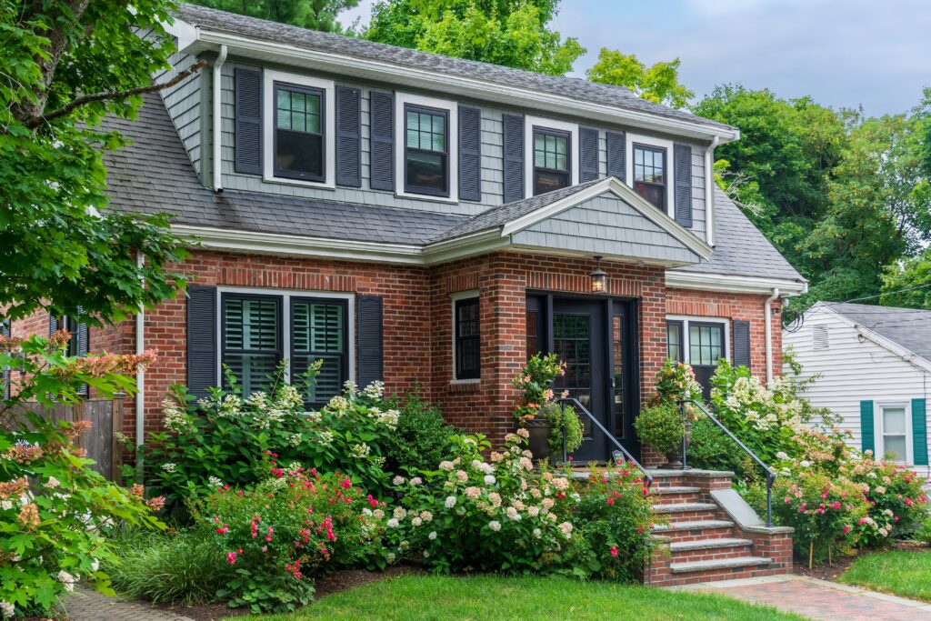 A two-story brick house with black shutters, flower gardens, and green shrubs. Steps with black railings lead to a dark front door. Trees surround the house under a partly cloudy sky, enhanced by expert Madison WI landscaping services.