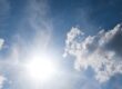 Bright summer sun shining through scattered clouds in a blue sky, representing the heat and dry conditions that stress trees in August.